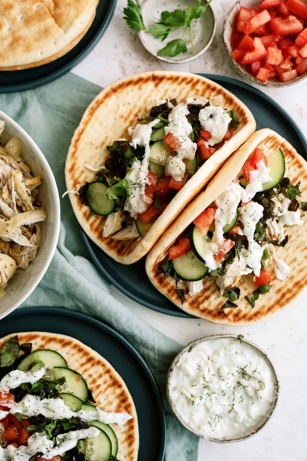Two pita sandwiches filled with sliced cucumbers, lettuce, tomatoes, shredded chicken, and drizzled with white sauce, displayed alongside bowls of tzatziki, tomatoes, and herbs.