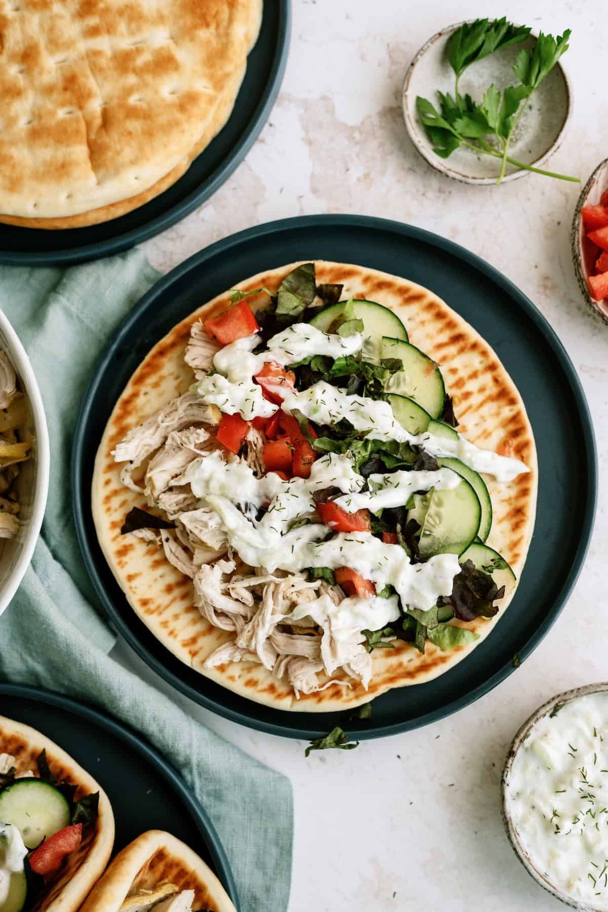 Flatbread topped with shredded chicken, sliced cucumber, lettuce, tomato, and a drizzle of white sauce, served on a dark plate with garnishes and extra sauce nearby.
