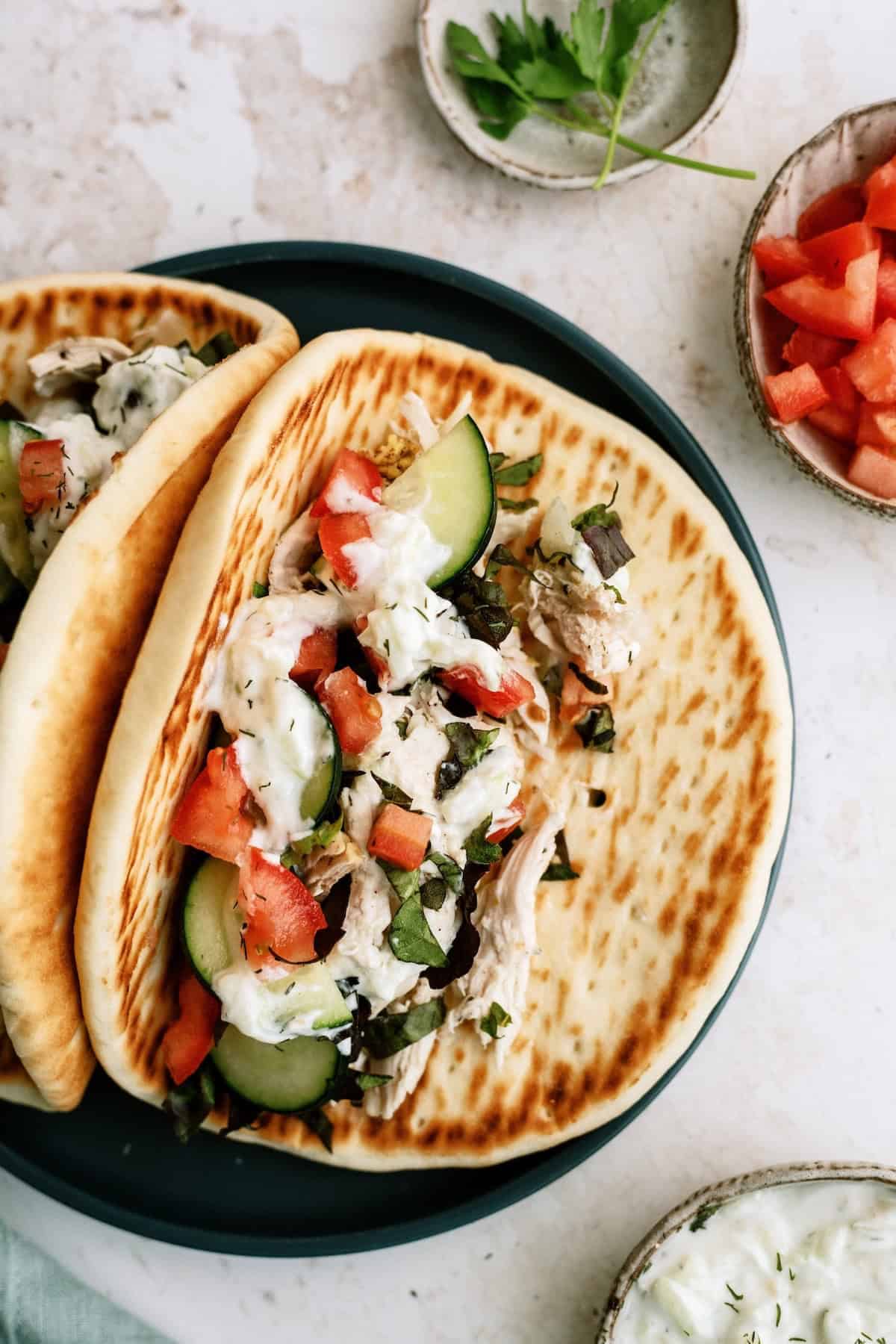 Two pita breads filled with sliced cucumber, chopped tomato, shredded chicken, and tzatziki sauce are served on a plate, with extra diced tomatoes and herbs in small bowls nearby.