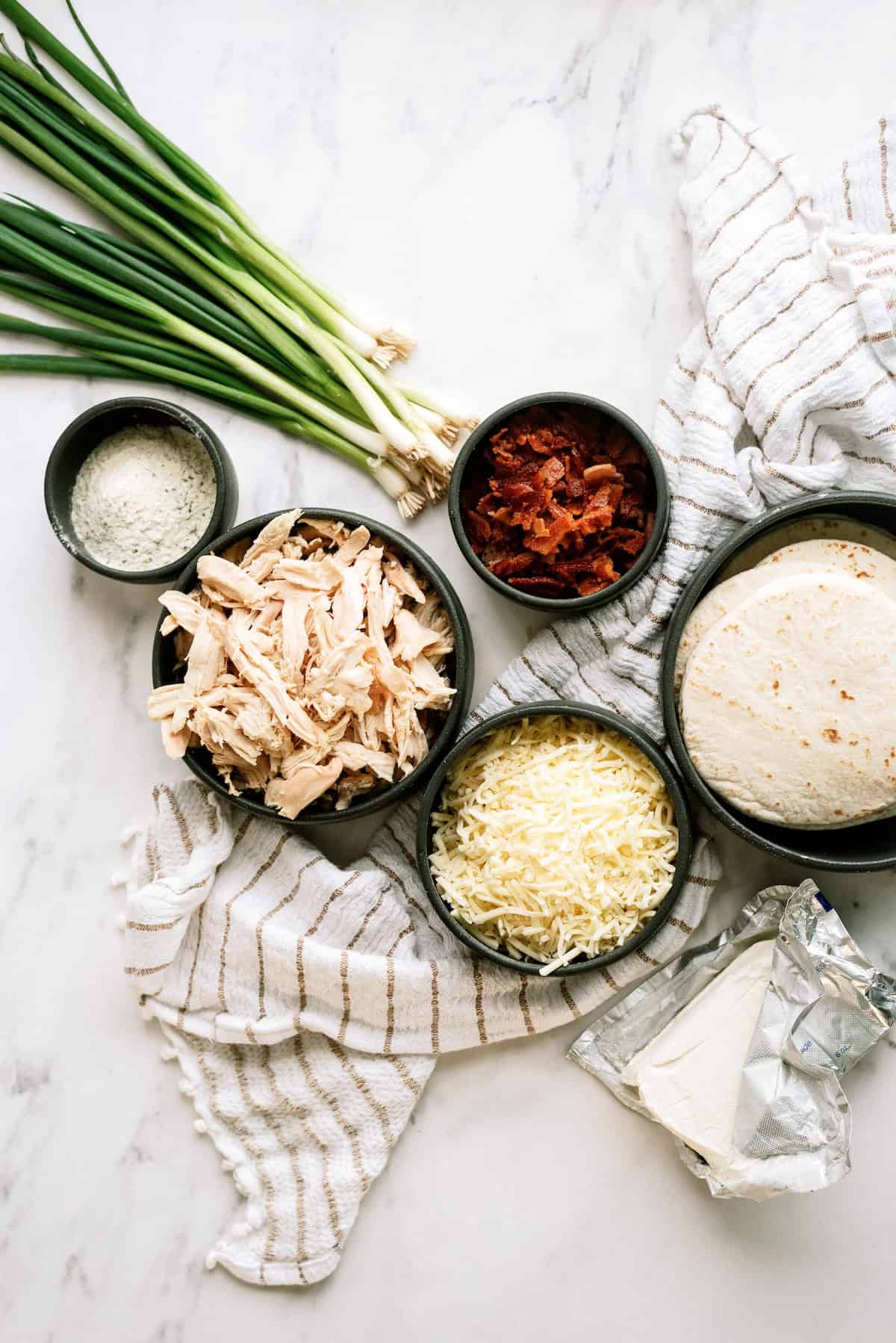 Flat lay of ingredients on a marble surface: shredded chicken, grated cheese, chopped bacon, green onions, tortillas, cream cheese, and a bowl of grated parmesan.