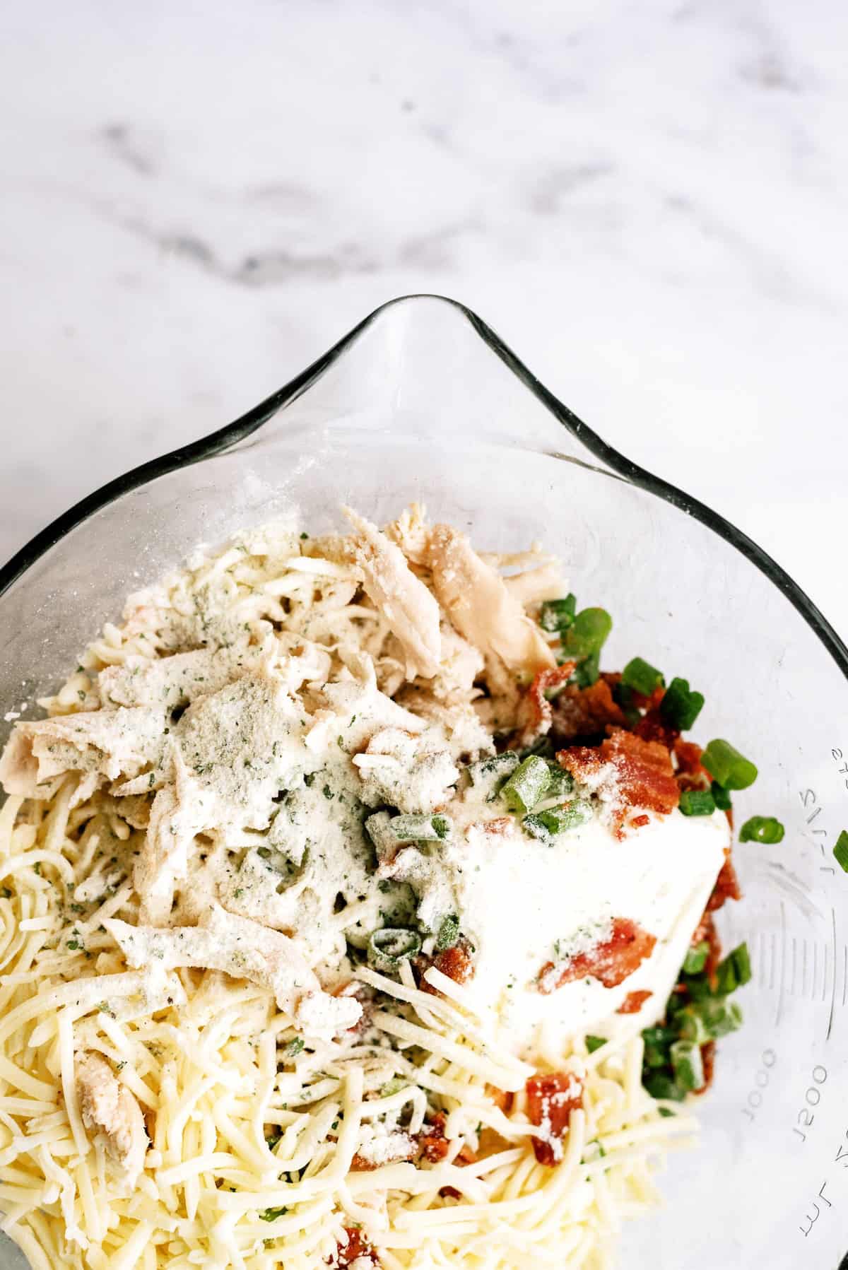 A glass mixing bowl filled with shredded cheese, chopped green onions, shredded chicken, tomato sauce, cream cheese, and powdered seasoning on a white surface.