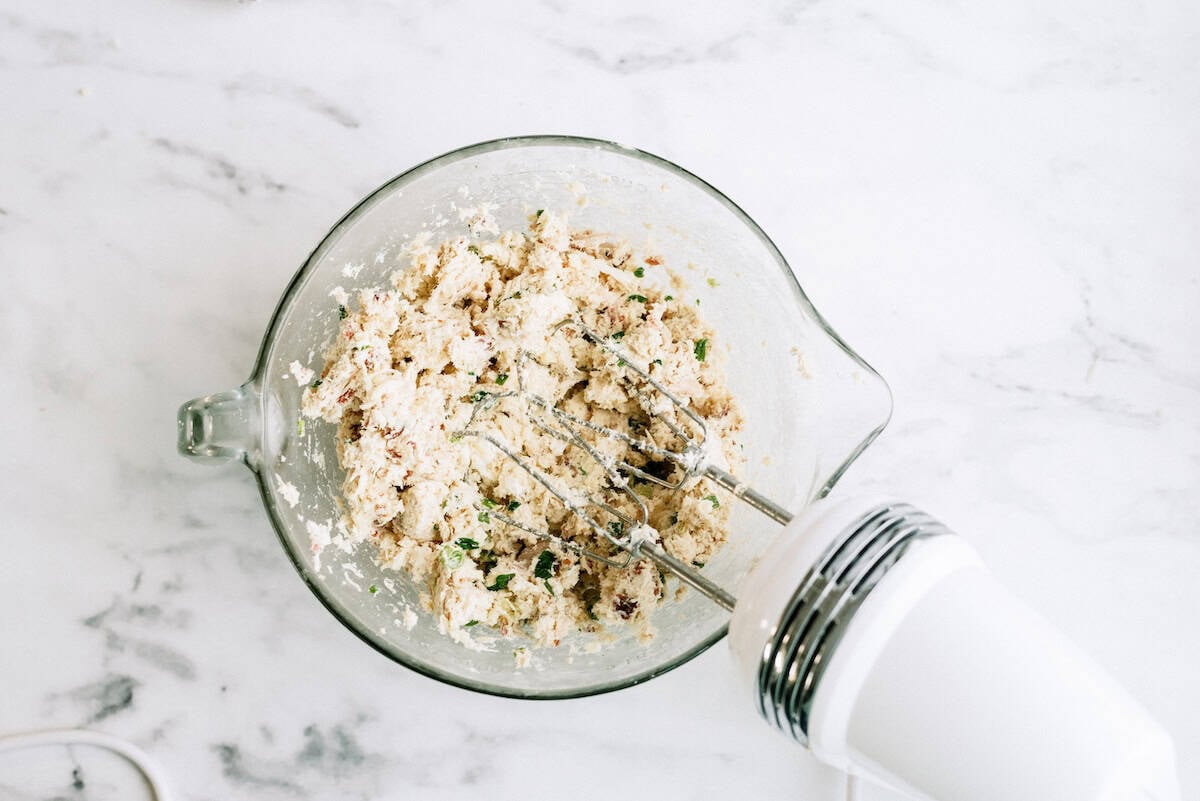 A glass mixing bowl with a mixture of ingredients and chopped herbs being blended by a handheld electric mixer on a marble countertop.