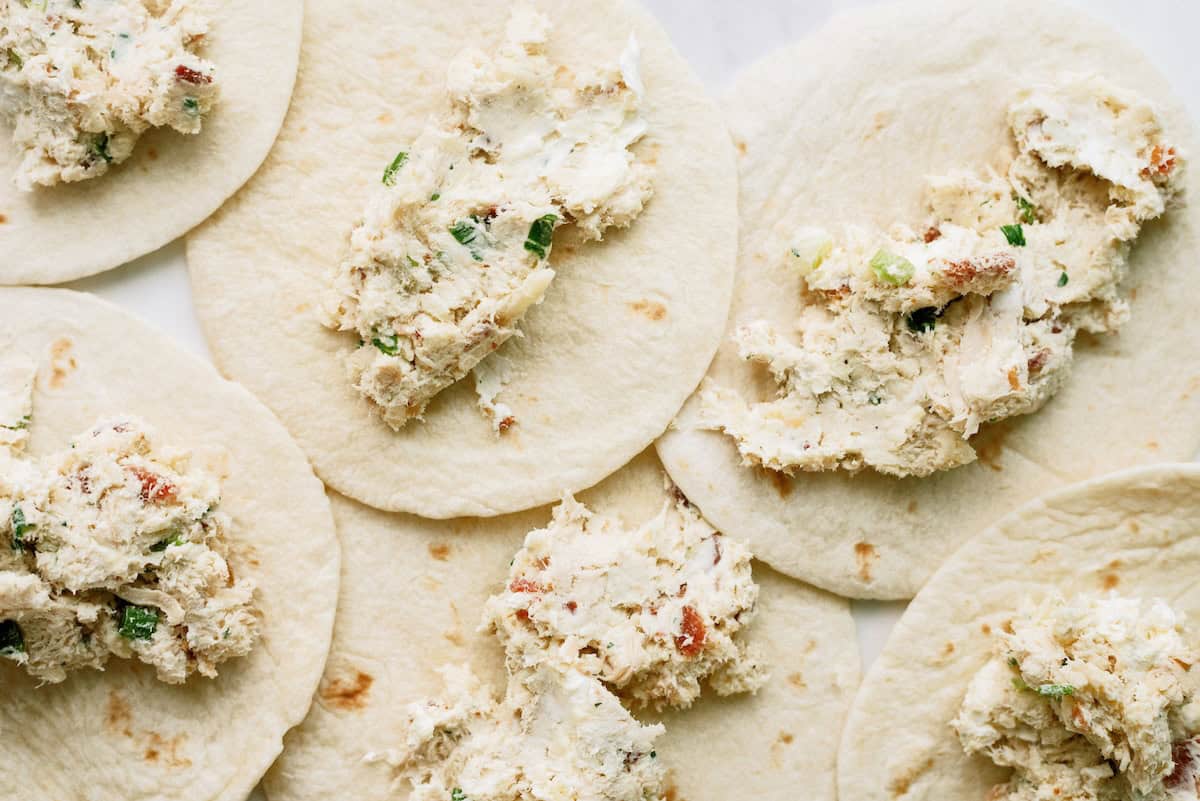 Several flour tortillas laid out flat with dollops of a creamy, chunky filling containing visible herbs and small bits of other ingredients.
