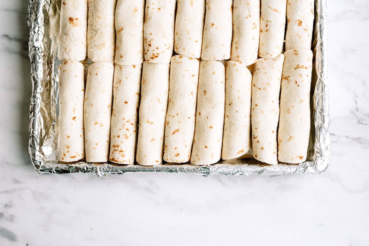 A foil-lined baking tray filled with neatly arranged, uncooked rolled tortillas on a white marble surface.