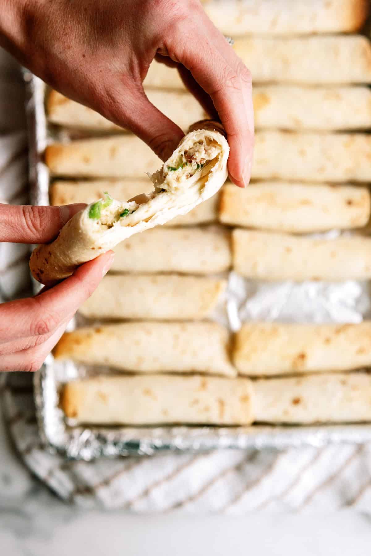 A hand holding a halved taquito filled with chicken and green vegetables over a tray of neatly arranged baked taquitos.