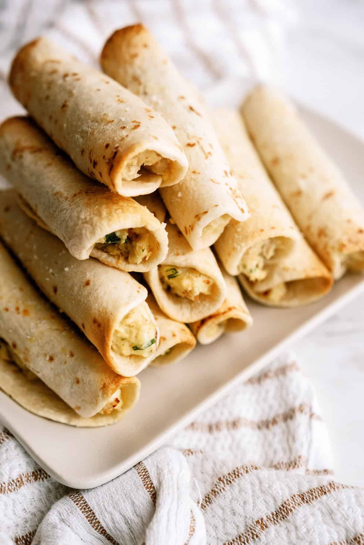 A plate stacked with several rolled tortillas filled with a light mixture, placed on a white and beige striped cloth.