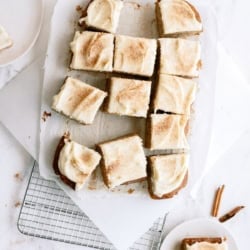 Overhead view of frosted square cake slices on parchment paper atop a cooling rack, with two small plates holding individual pieces and cinnamon sticks nearby.