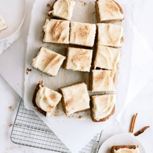 Overhead view of frosted square cake slices on parchment paper atop a cooling rack, with two small plates holding individual pieces and cinnamon sticks nearby.