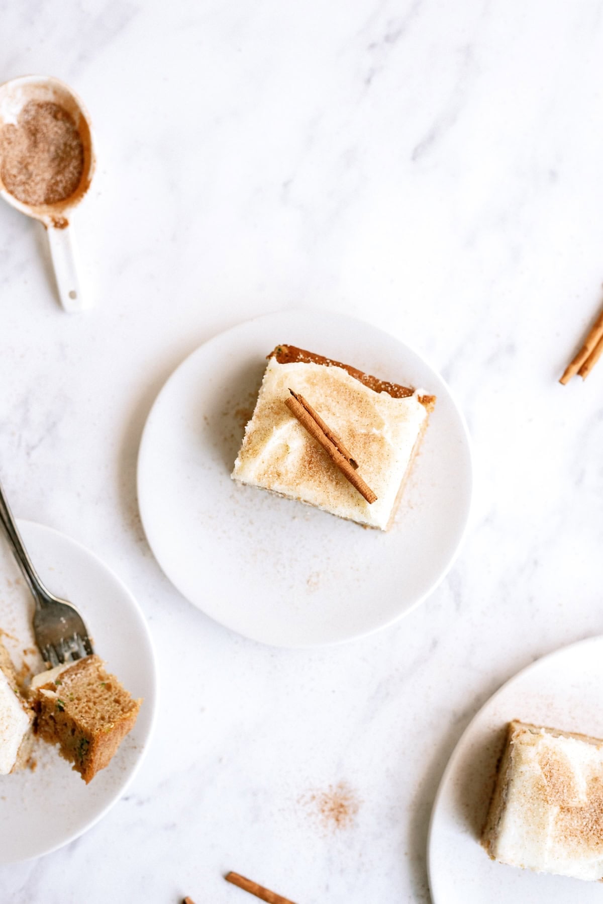 A square piece of frosted cake topped with a cinnamon stick on a white plate, with another slice and cinnamon nearby on a marble surface.