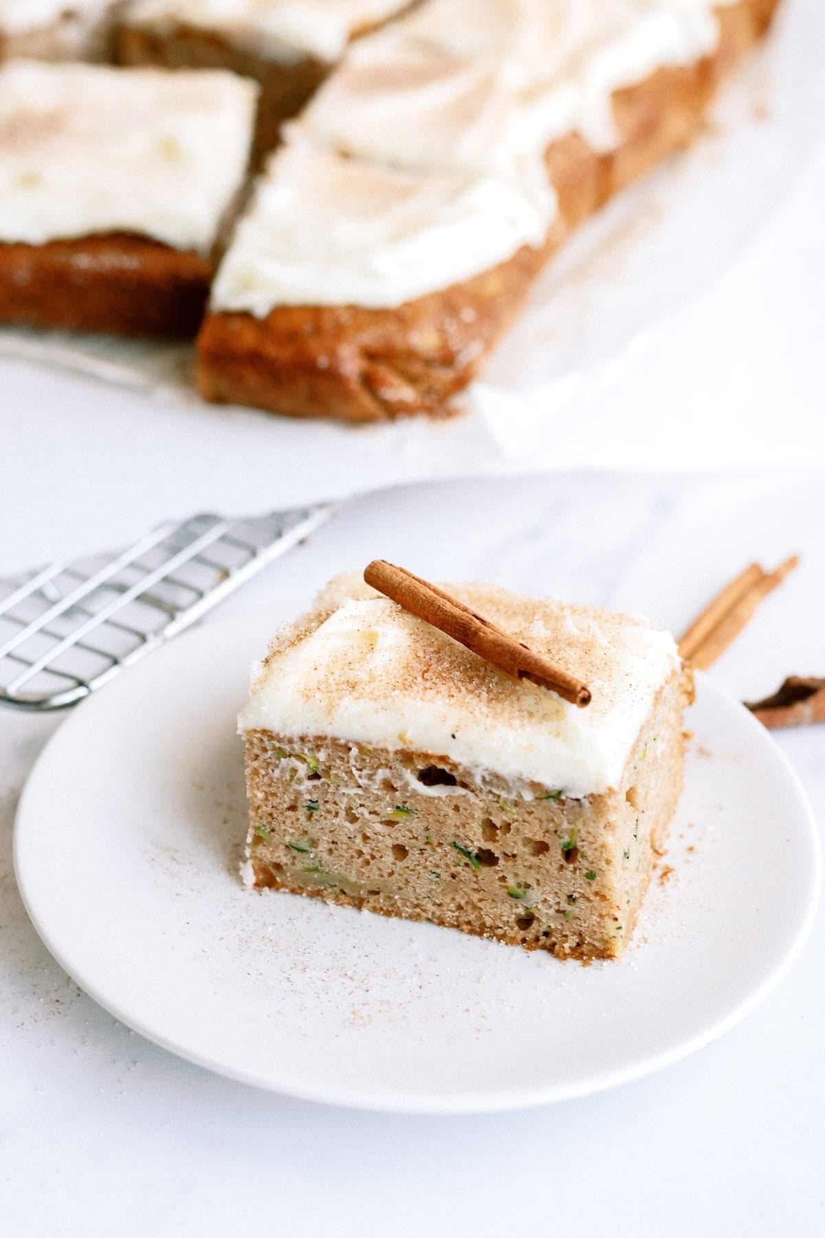 A square piece of frosted cake topped with a cinnamon stick is on a white plate, with more cake pieces visible in the background.