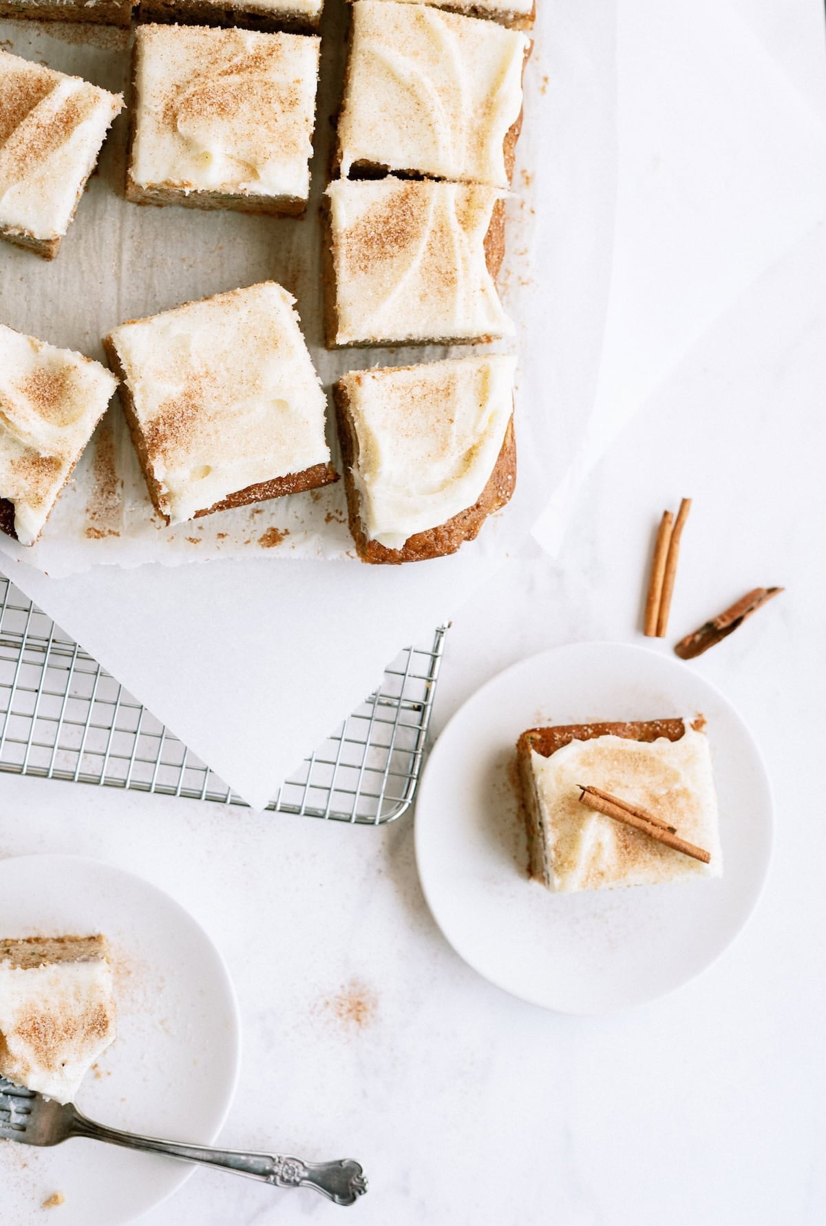 Sliced frosted cake squares on a cooling rack and plate, topped with cinnamon, with a fork and cinnamon sticks on a white surface.