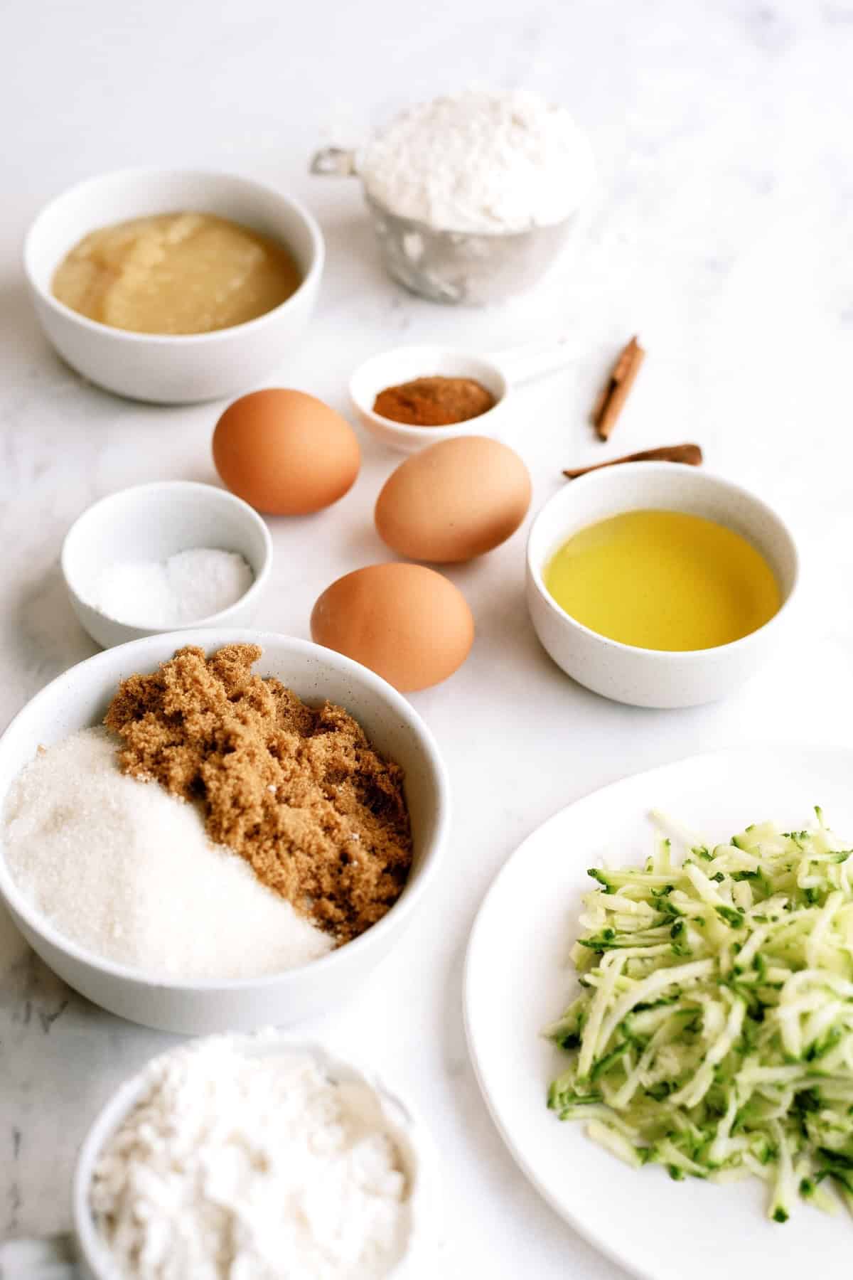 Various baking ingredients including eggs, shredded zucchini, brown and white sugar, flour, oil, applesauce, salt, cinnamon, and baking powder arranged on a white surface.