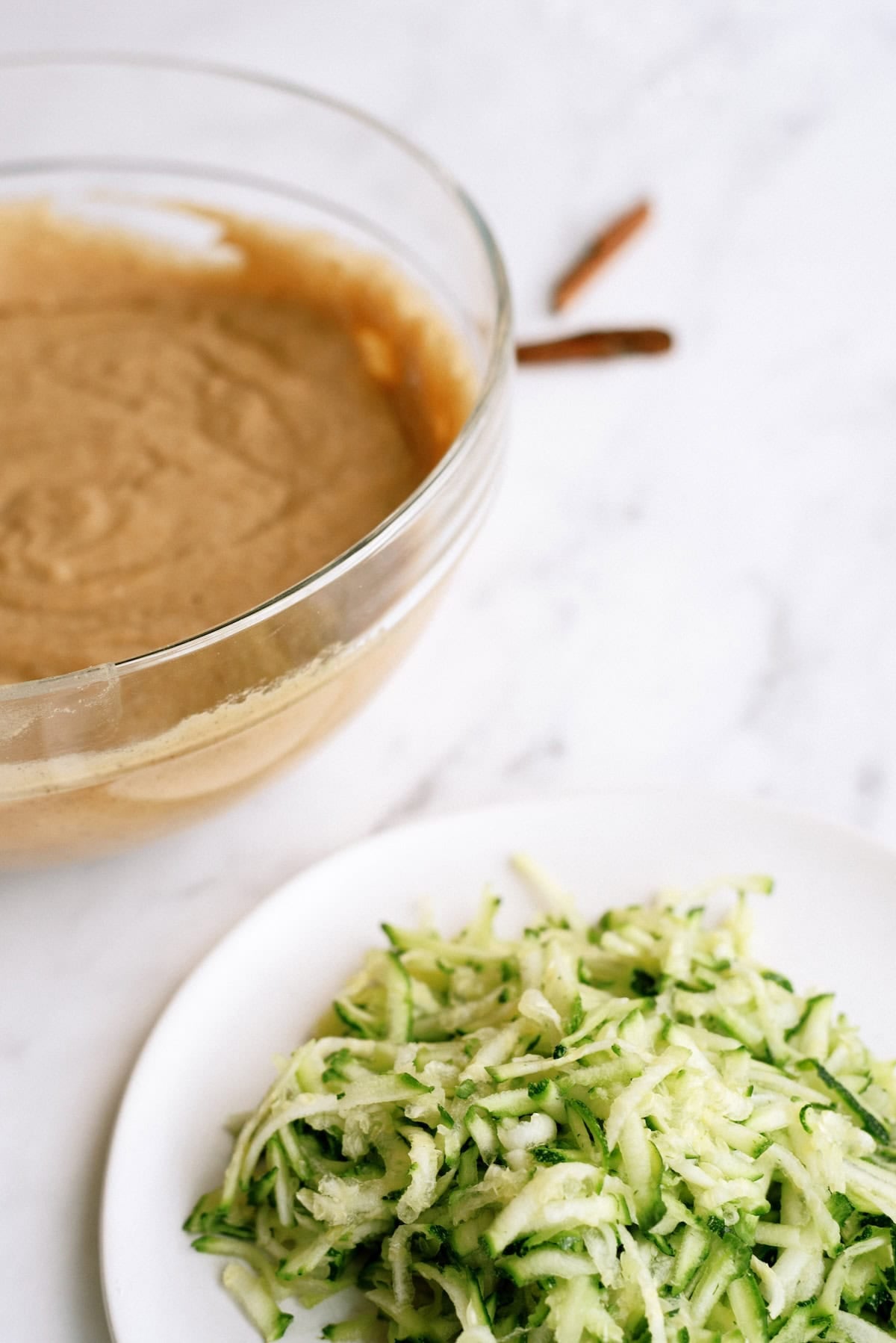 A glass bowl of batter sits next to a plate of shredded zucchini on a white surface.
