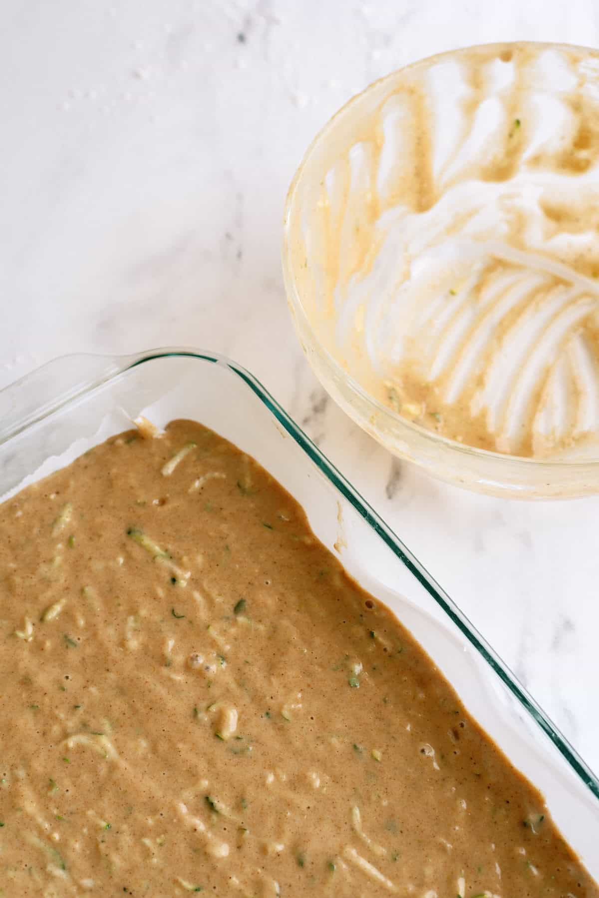 A glass baking dish filled with cake batter sits on a marble surface next to an empty, batter-smeared mixing bowl.