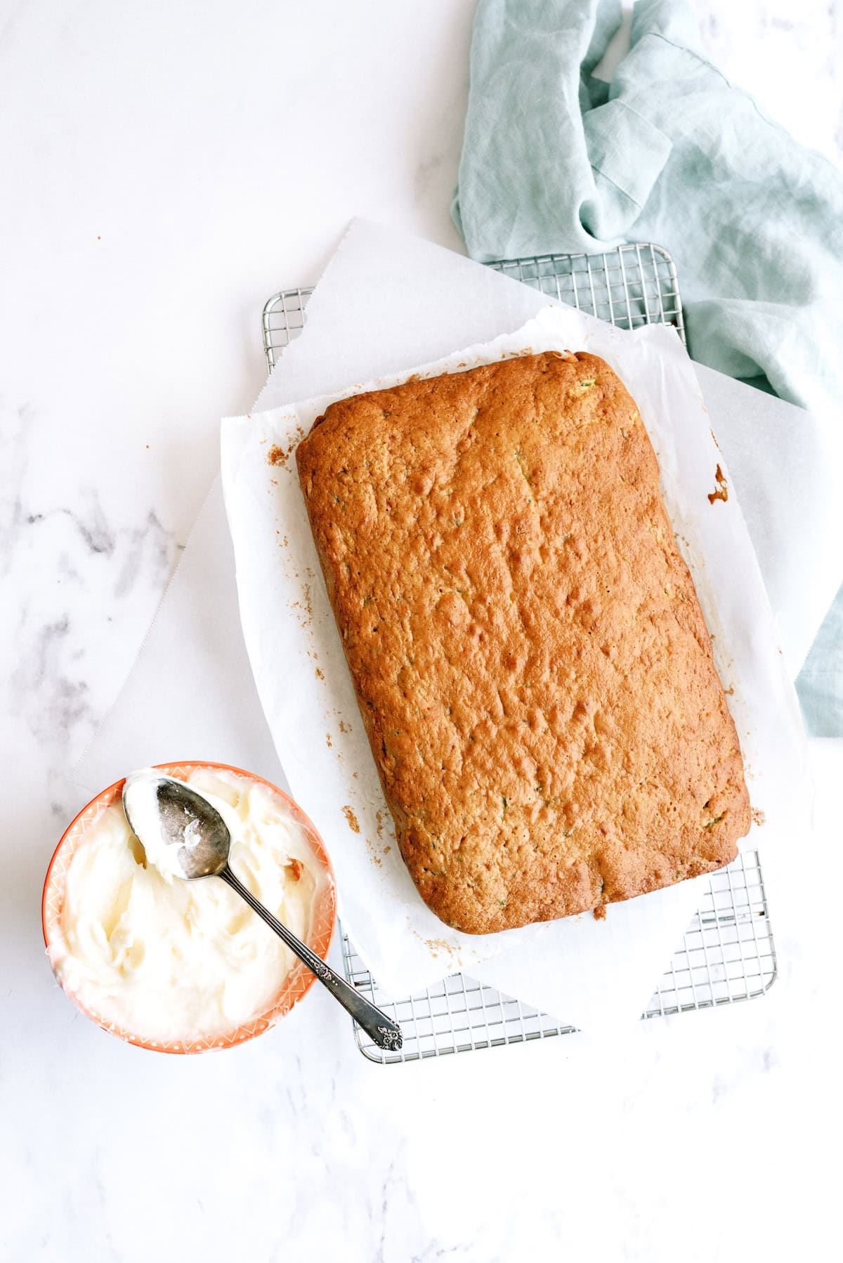 A loaf of banana bread on parchment paper atop a cooling rack, with a bowl of frosting and a spoon nearby on a marble surface. A folded light green cloth is in the corner.