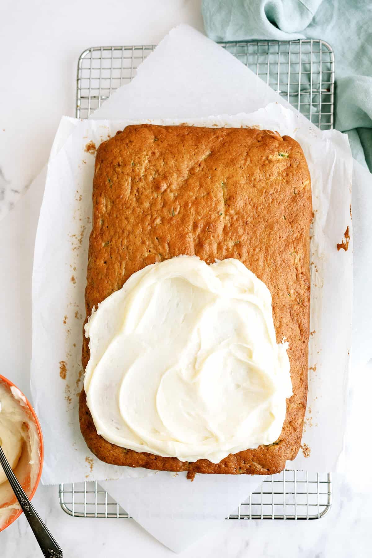 A rectangular cake on parchment paper is partially frosted with white icing, resting on a cooling rack with a bowl of icing and a towel nearby.