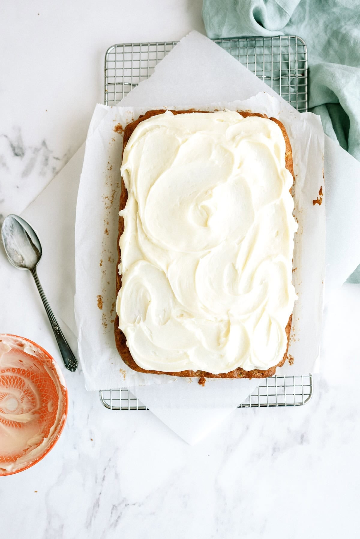 A rectangular cake with white frosting sits on parchment paper atop a cooling rack, with a spoon and an empty bowl nearby on a marble surface.