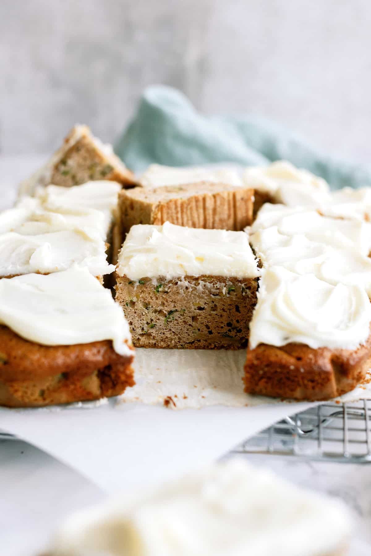 Slices of zucchini cake with white frosting are arranged on parchment paper, with one piece set upright to show the moist texture inside.