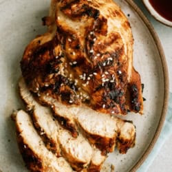 Sliced grilled chicken breast topped with sesame seeds on a round ceramic plate, accompanied by dipping sauces in small bowls.