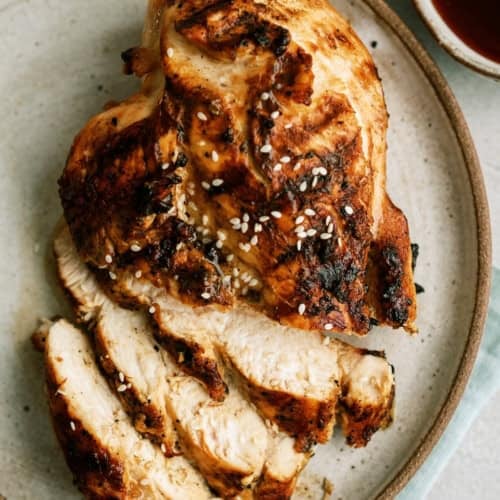 Sliced grilled chicken breast topped with sesame seeds on a round ceramic plate, accompanied by dipping sauces in small bowls.