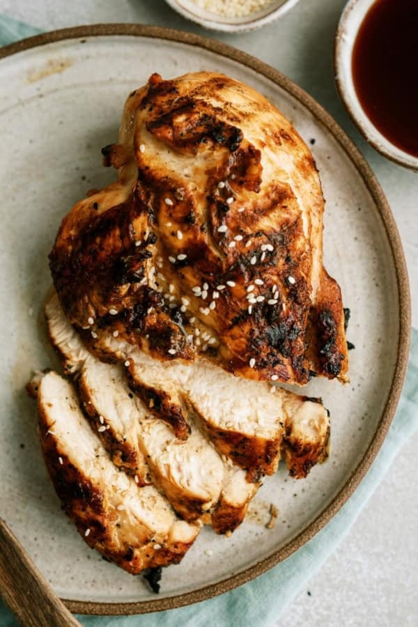 Sliced grilled chicken breast topped with sesame seeds on a round ceramic plate, accompanied by dipping sauces in small bowls.