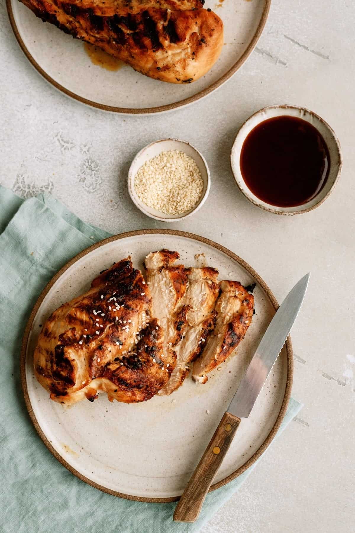 Grilled chicken breast on a plate with a knife, garnished with sesame seeds, next to small bowls of sesame seeds and sauce.