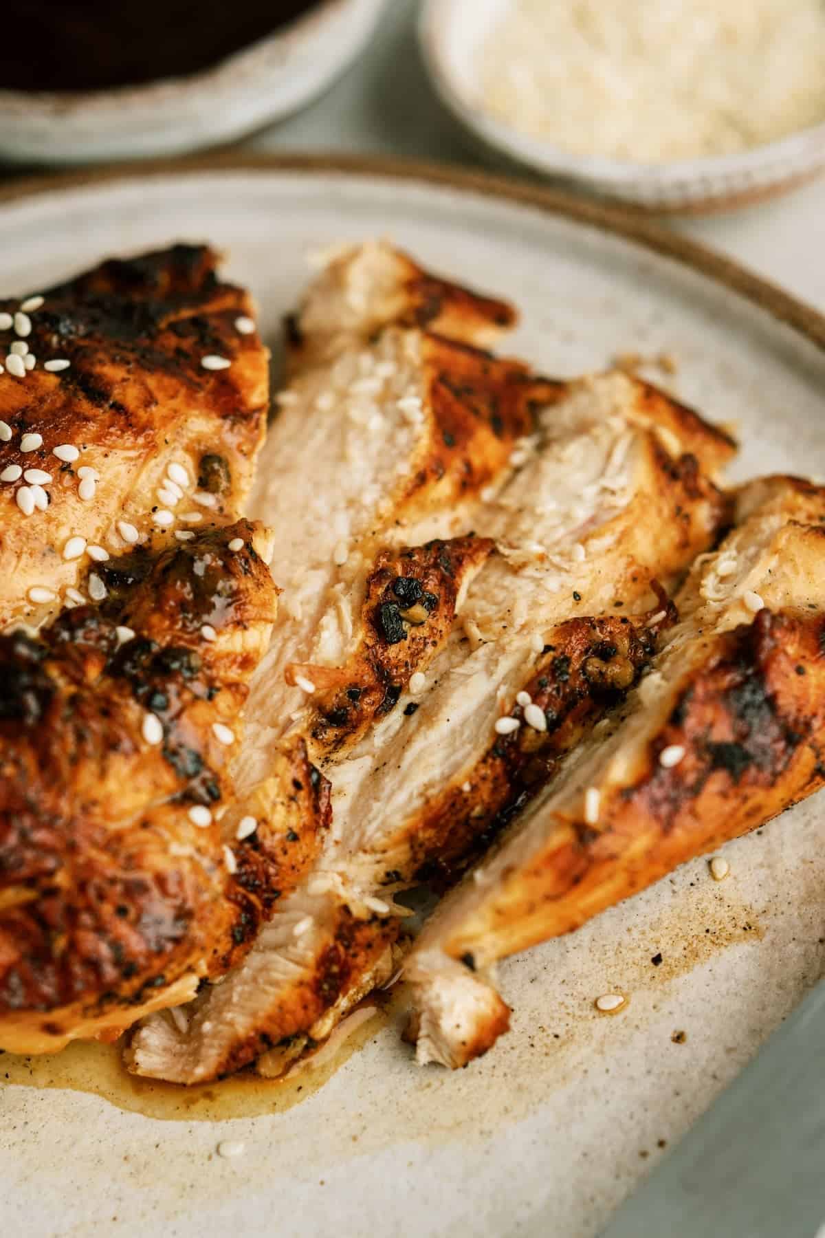 Sliced grilled chicken breast garnished with sesame seeds on a beige plate, with bowls of sauce and grated cheese in the background.
