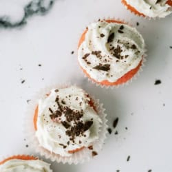 Four cupcakes with white frosting and chocolate shavings on top are arranged in a line on a white surface.