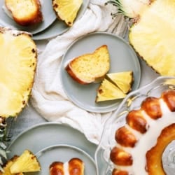 Sliced pineapple and pieces of glazed bundt cake are arranged on gray plates with a cake on a glass stand.