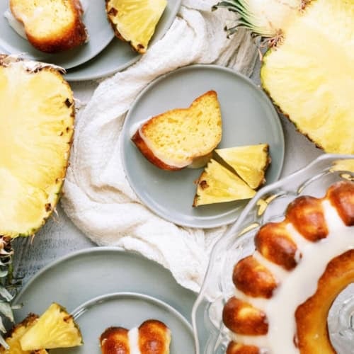Sliced pineapple and pieces of glazed bundt cake are arranged on gray plates with a cake on a glass stand.
