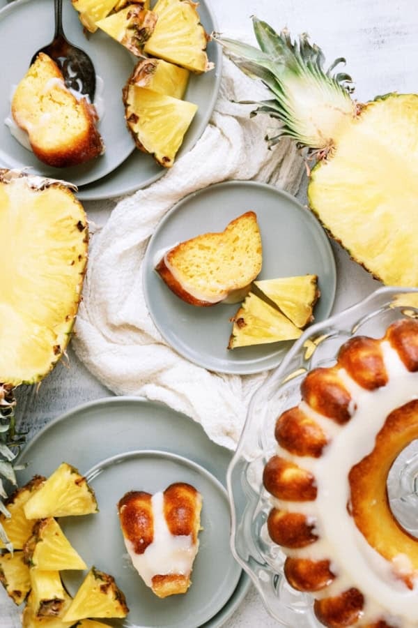 Sliced pineapple and pieces of glazed bundt cake are arranged on gray plates with a cake on a glass stand.