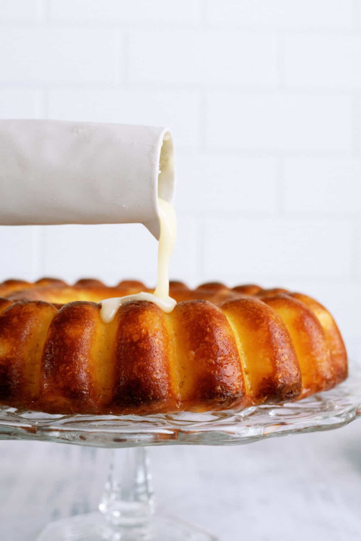 A white pitcher pours glaze onto a round bundt cake displayed on a glass cake stand against a white tiled background.