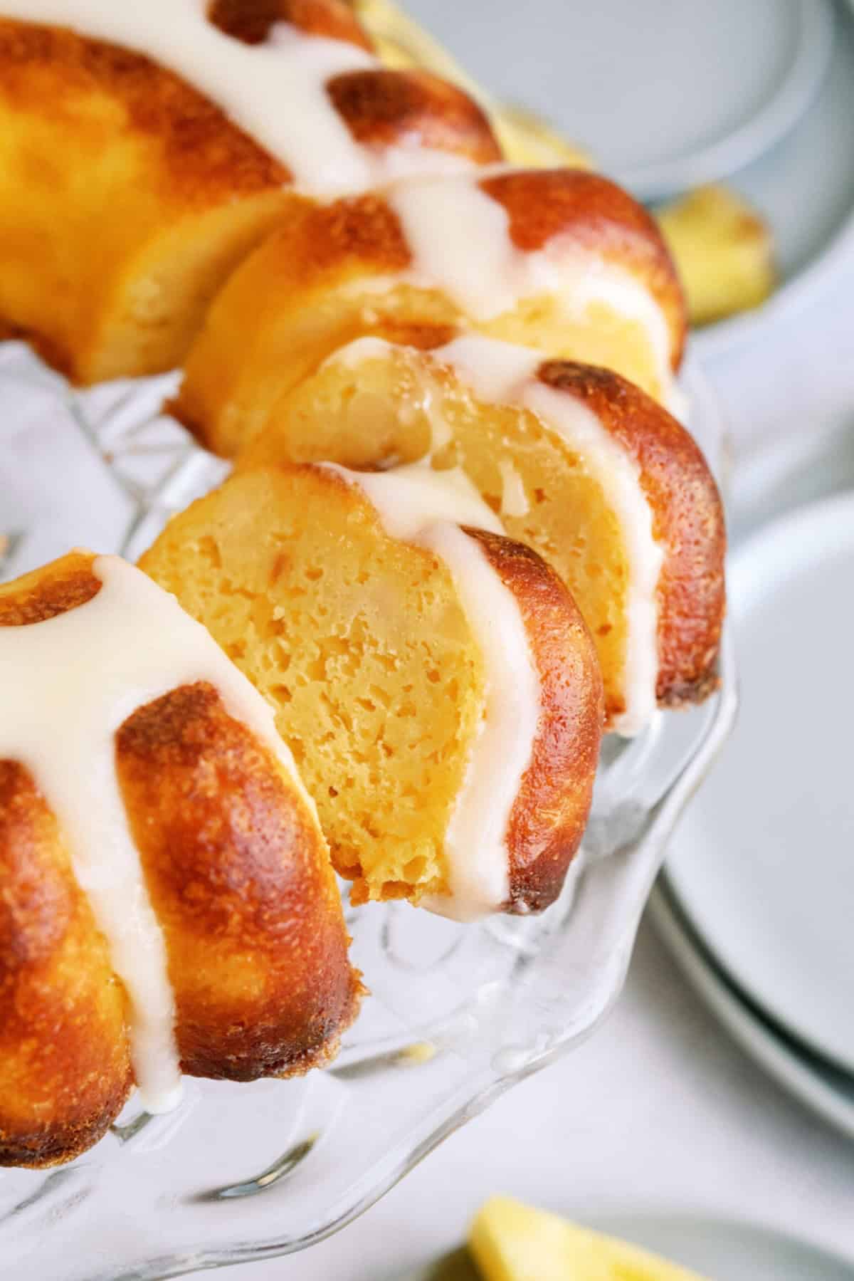 A close-up of a sliced bundt cake with a golden crust and white glaze drizzled on top, displayed on a glass cake stand.