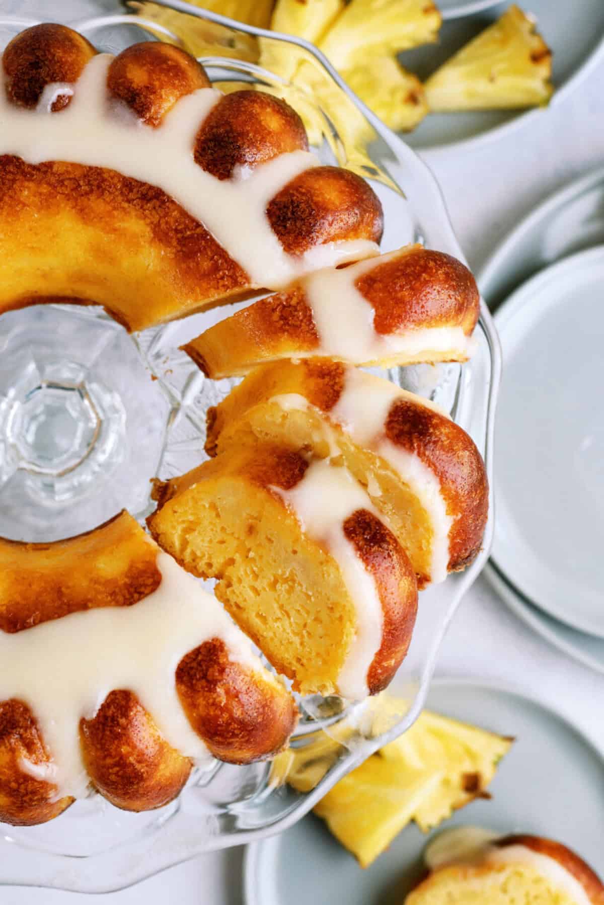 A sliced bundt cake with white icing on a glass cake stand, surrounded by pineapple slices and plates.