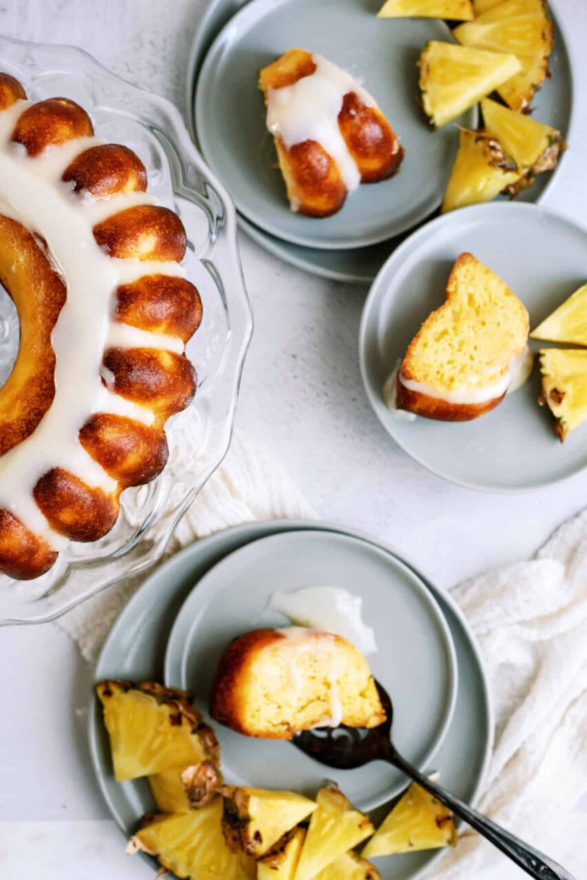 A glazed bundt cake on a glass stand, with slices served on gray plates alongside fresh pineapple wedges.