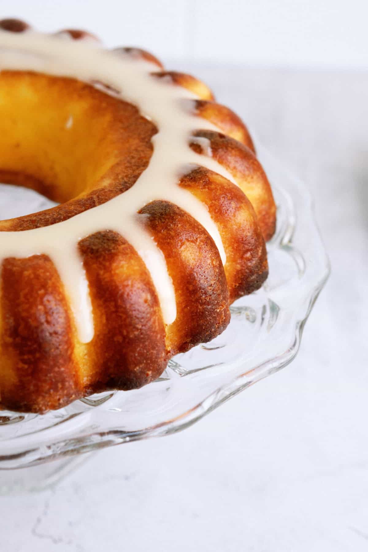 A bundt cake with a light glaze drizzled on top, displayed on a clear glass cake stand.