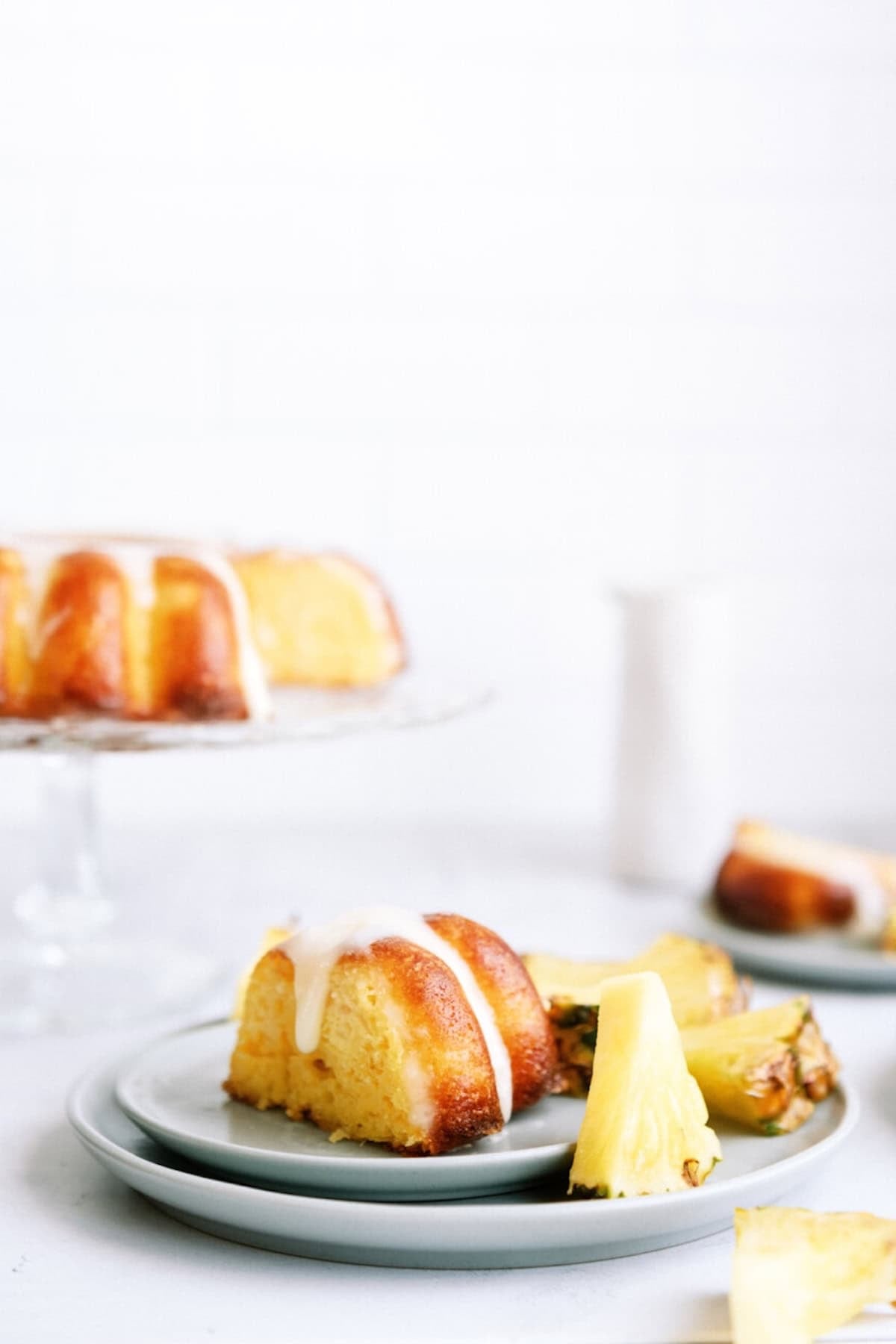 A slice of bundt cake with white icing and pineapple pieces on a plate, with more cake in the background.