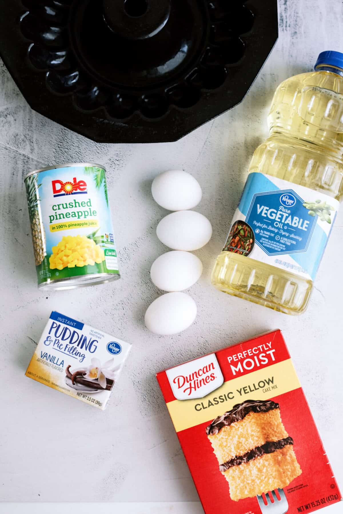 Ingredients for cake on a white surface: a Bundt pan, vegetable oil, four eggs, crushed pineapple, vanilla pudding mix, and classic yellow cake mix.