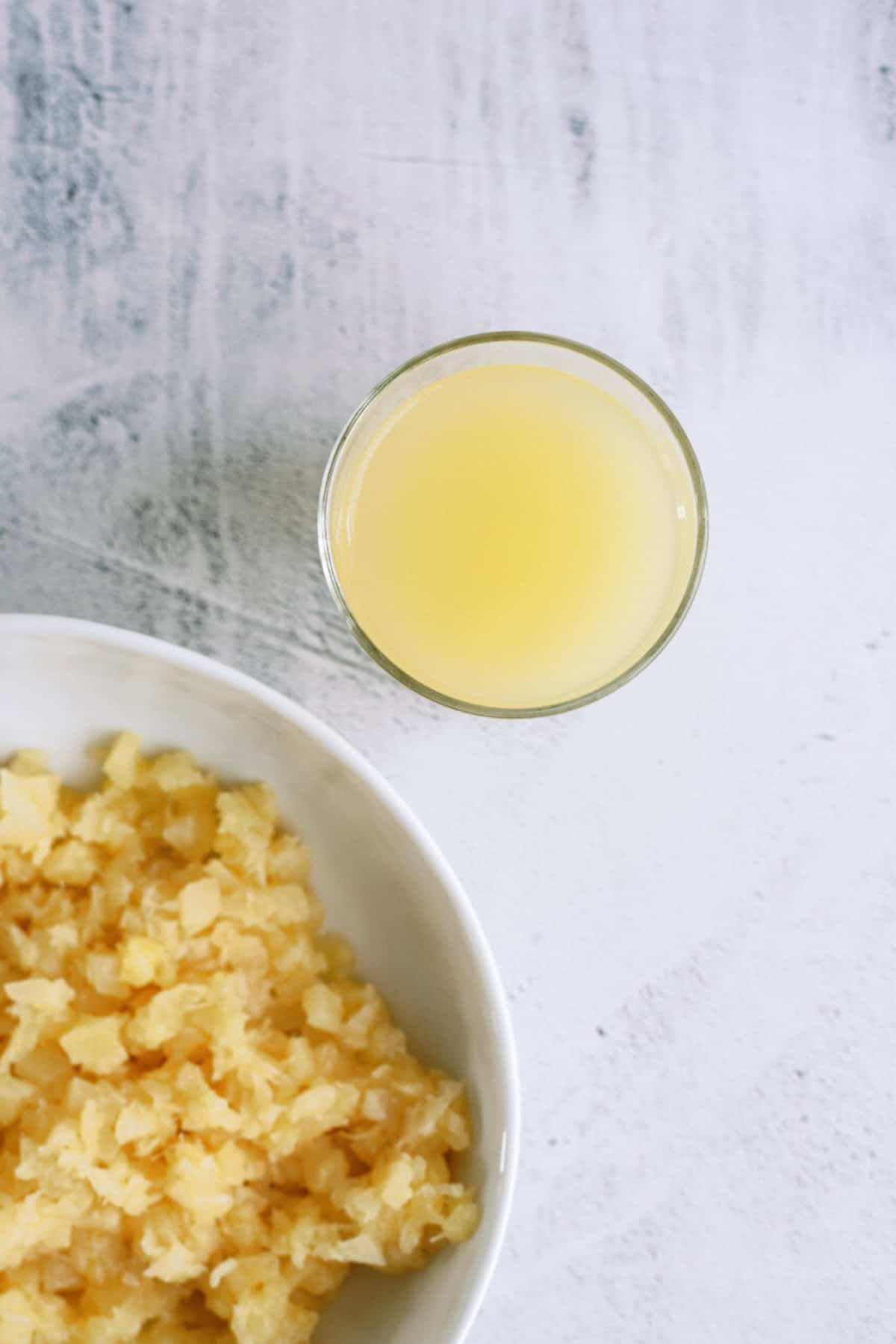 A glass of pineapple juice next to a bowl of chopped pineapple on a white surface.
