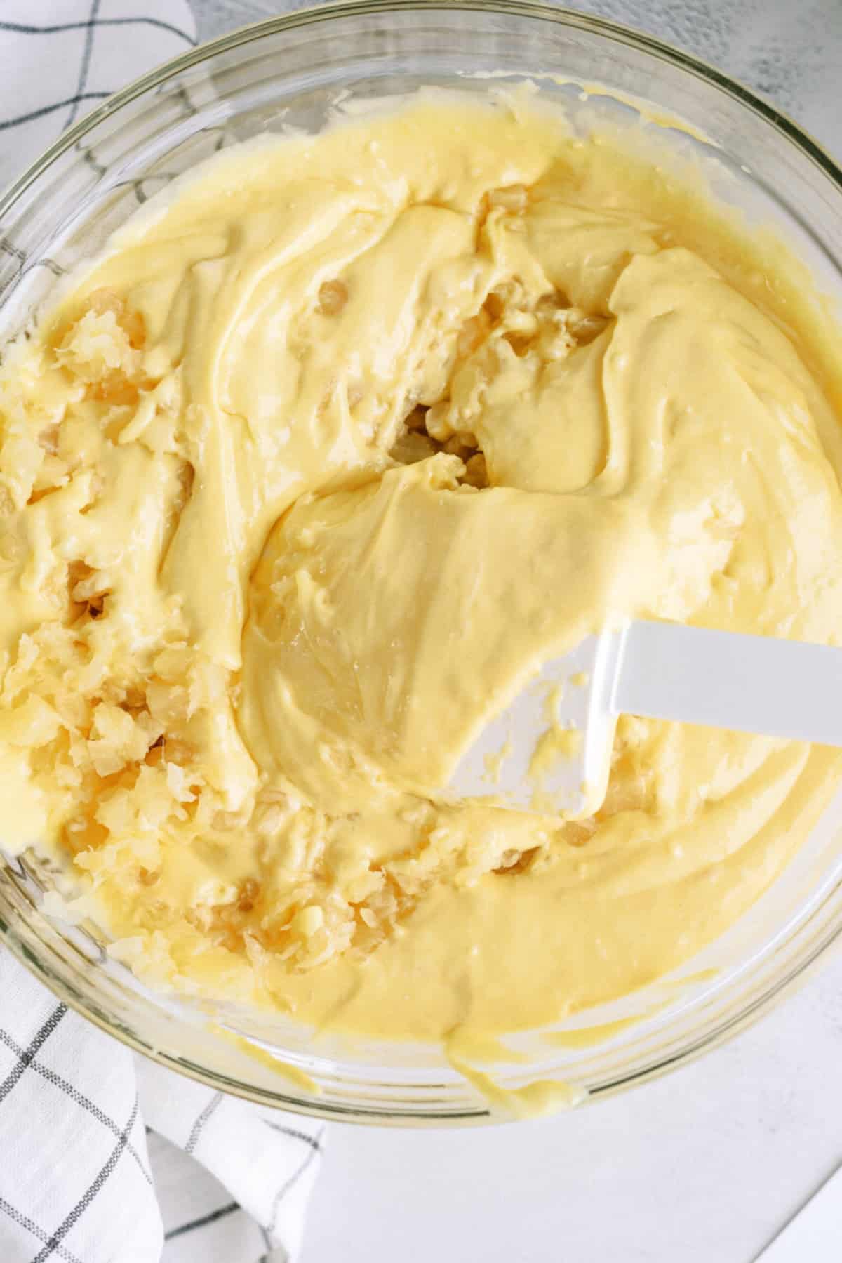 A glass bowl of yellow cake batter being mixed with crushed pineapple using a white spatula, next to a white and black checkered cloth.