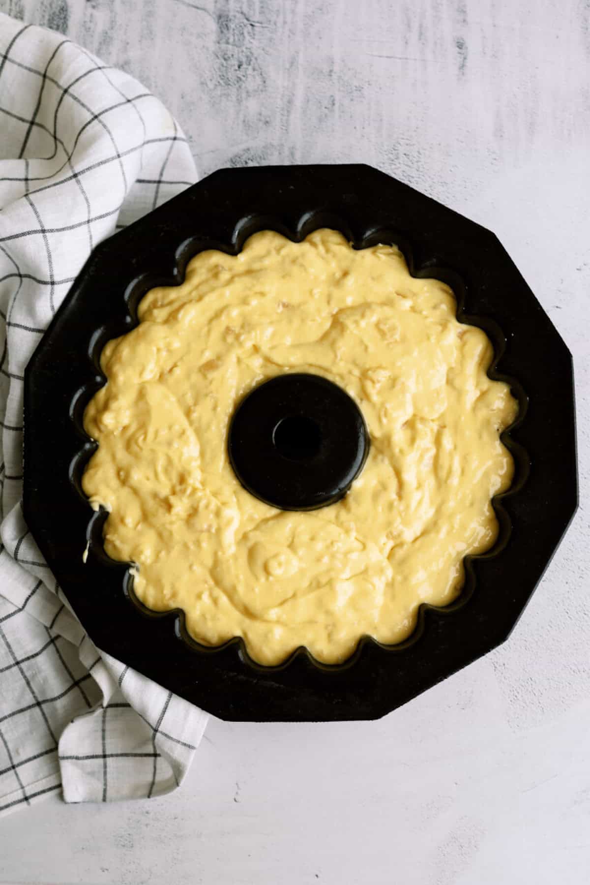 A black bundt cake pan filled with yellow cake batter sits on a white surface next to a white kitchen towel with a black grid pattern.