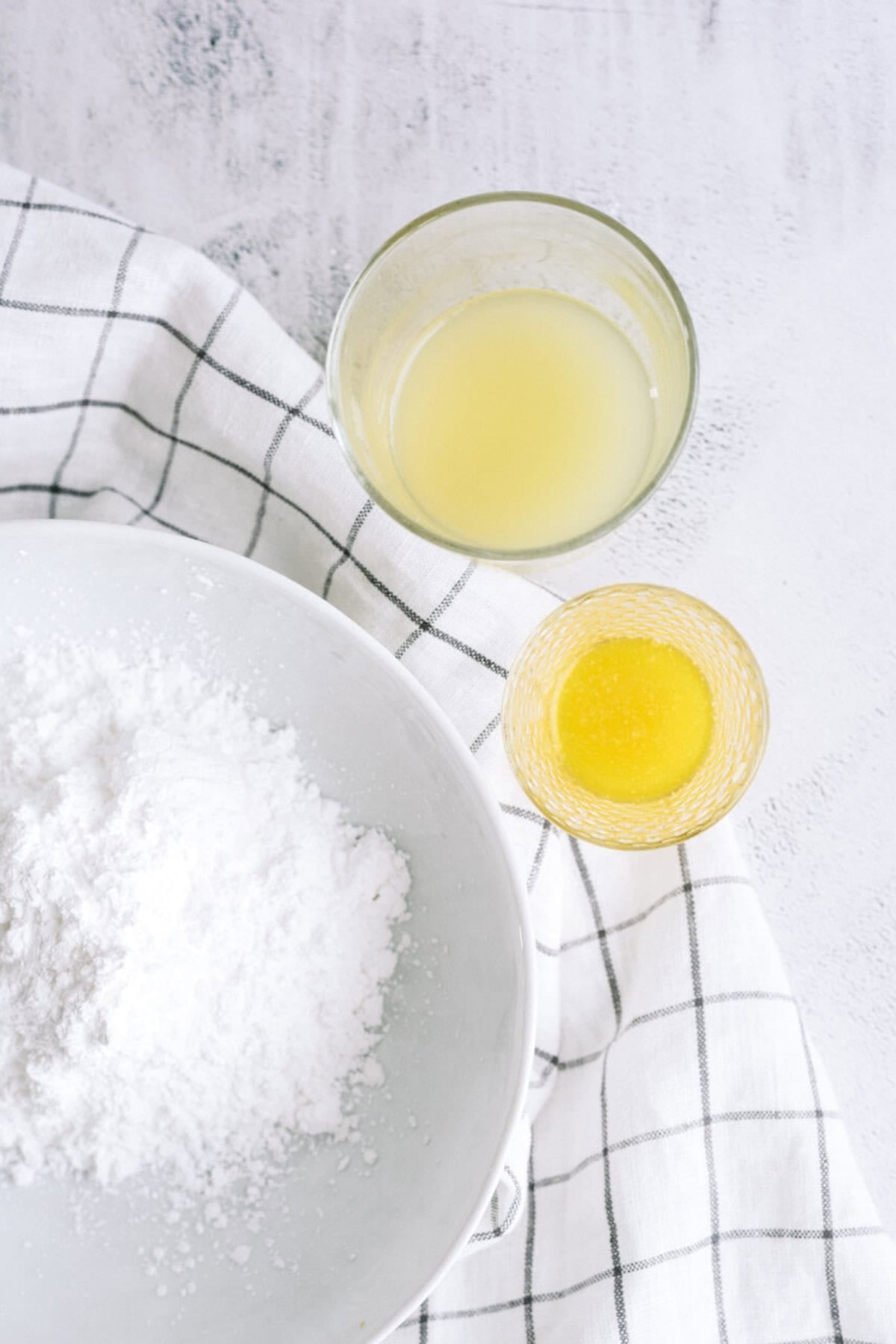 A bowl of powdered sugar next to a glass of lemon juice and a glass of melted butter on a white checkered cloth.