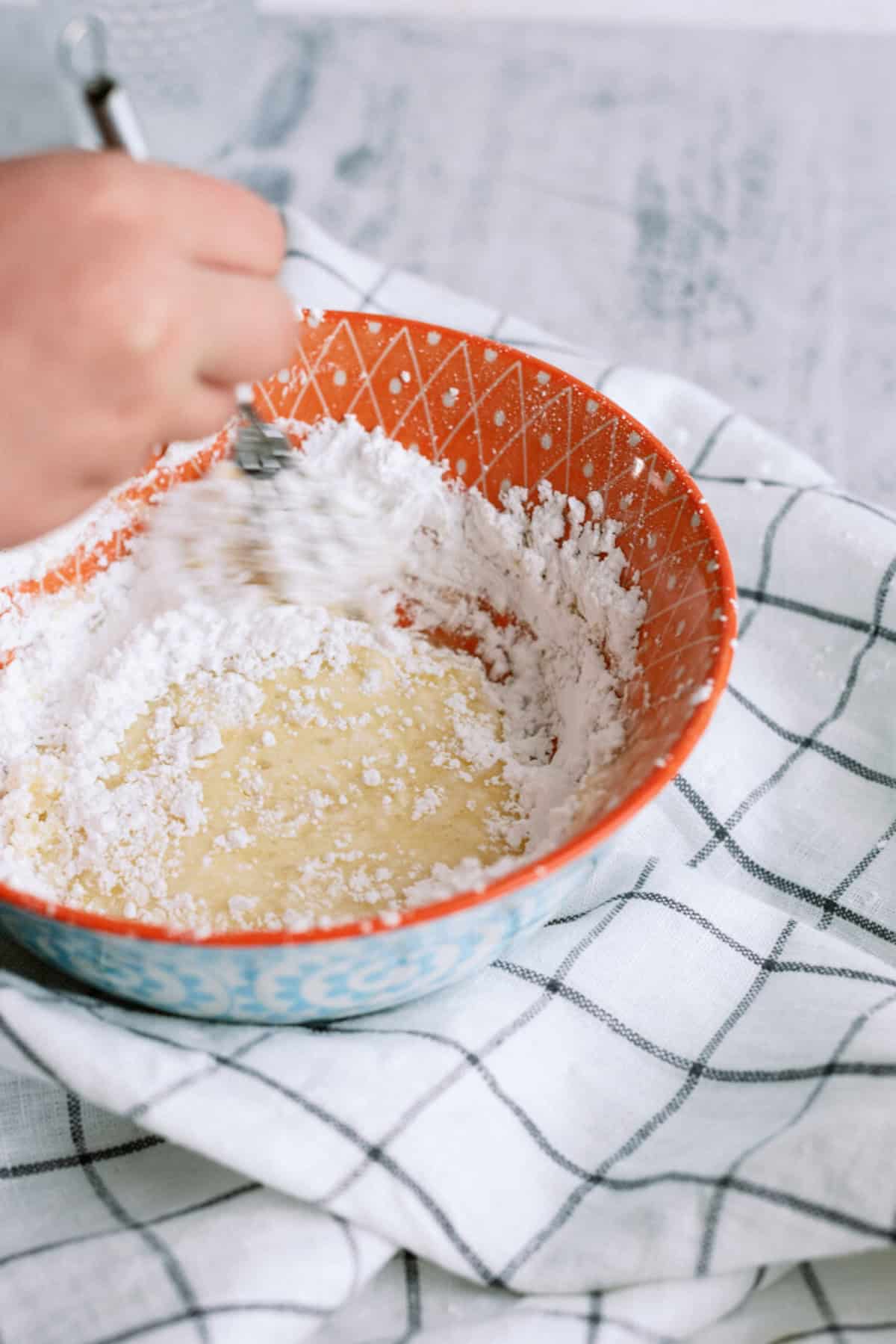 A hand mixing white powder and liquid in a patterned orange bowl with a whisk, placed on a checkered cloth.