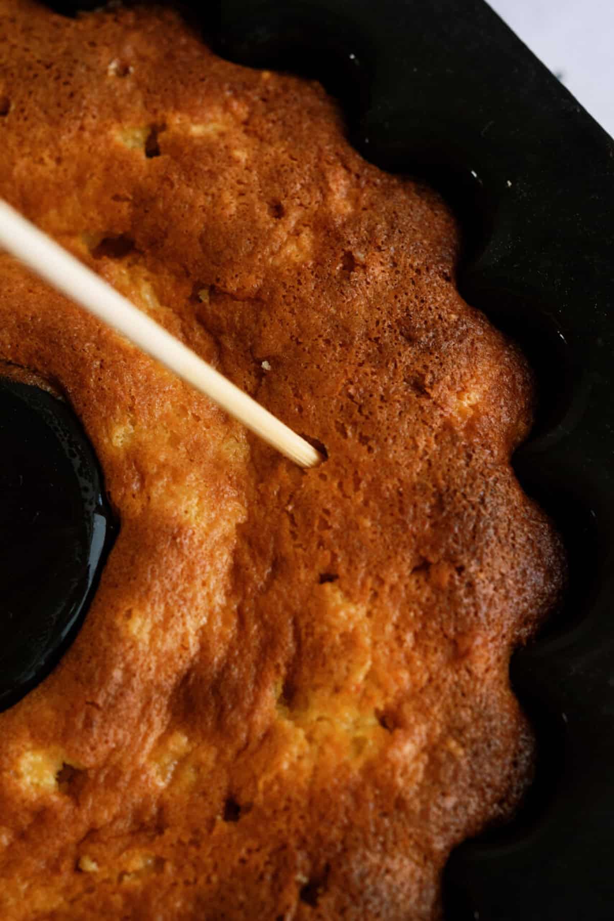 A wooden skewer is inserted into a golden brown ring-shaped cake to check for doneness.