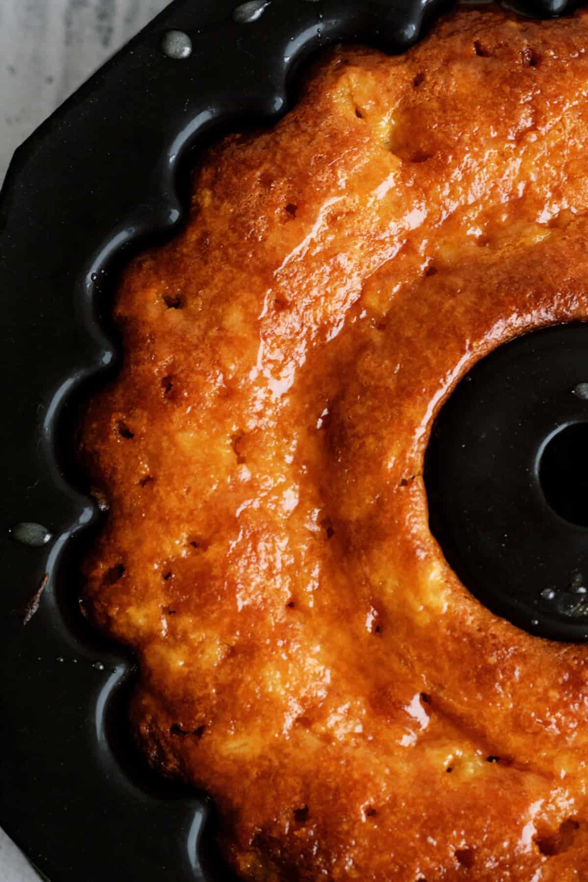 Close-up of a golden-brown bundt cake in a black bundt pan, showing a shiny, glazed surface and textured edges.