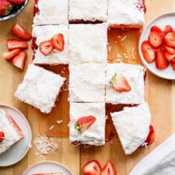 A rectangular coconut-topped cake is cut into squares, some garnished with sliced strawberries, and served on plates with extra strawberries on the side.