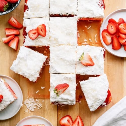 A rectangular coconut-topped cake is cut into squares, some garnished with sliced strawberries, and served on plates with extra strawberries on the side.