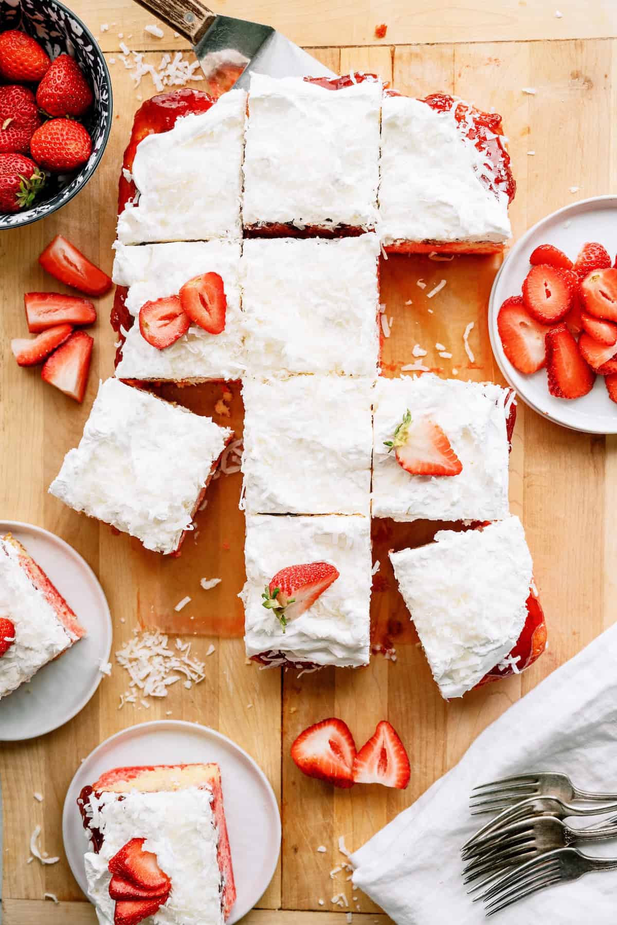 A rectangular coconut-topped cake is cut into squares, some garnished with sliced strawberries, and served on plates with extra strawberries on the side.