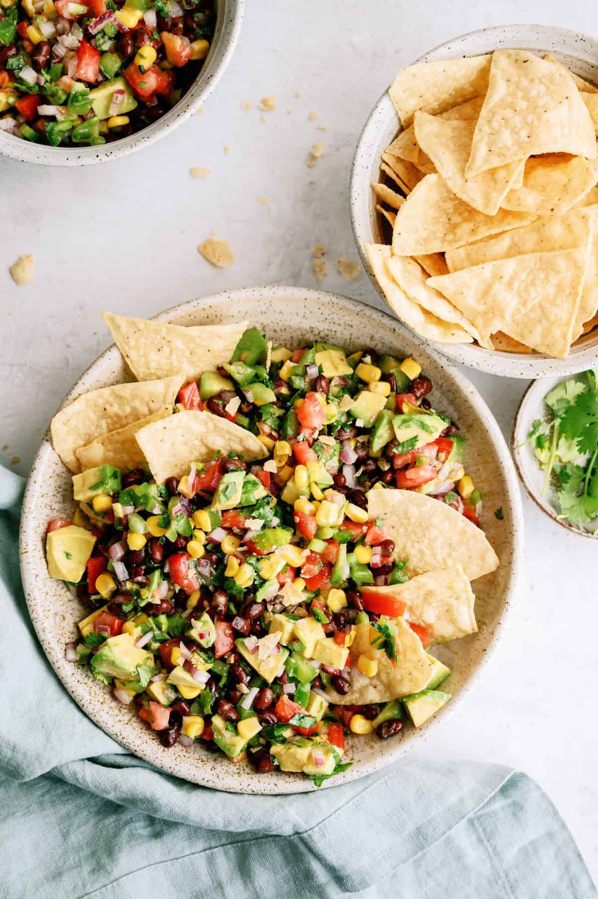 A bowl of corn, black bean, avocado, and tomato salsa served with tortilla chips, alongside bowls of chips and cilantro on a light surface.