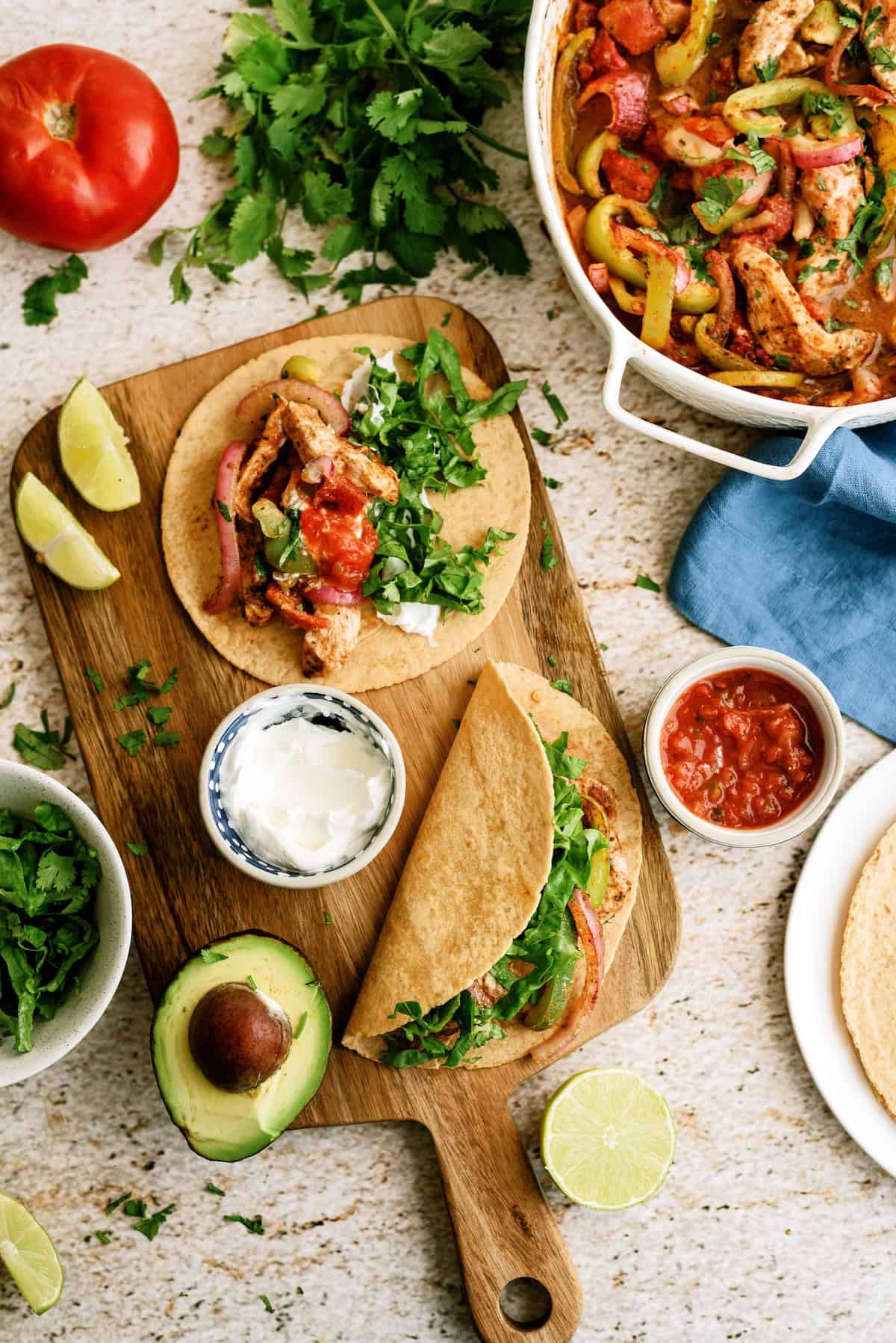 A wooden board with chicken fajitas, topped with lettuce and salsa, beside avocado, lime, cilantro, sour cream, and a pan of cooked chicken and peppers.